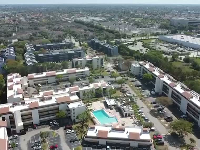Aerial over a low-rise coastal community — reflective roofs, pool decks and palm canopy.